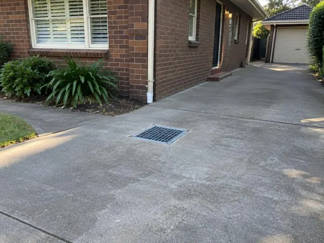 Concrete driveway with a square drain grate beside a brick house and garden in Redhead