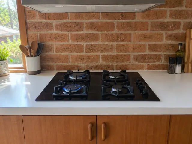 Black gas cooktop set into a white benchtop with brick splashback and timber cabinets in Whitebridge